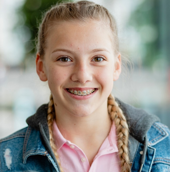 Young girl with braces smiling at camera, wearing denim jacket and pink shirt, indoor setting.