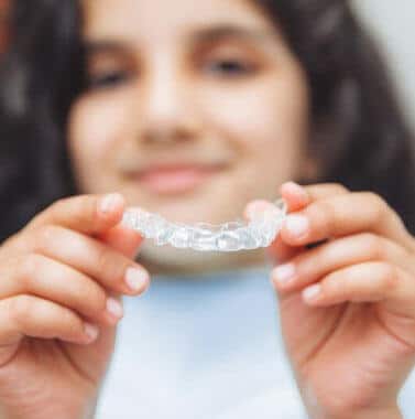 Young person holding clear orthodontic aligner, smiling at camera indoors.