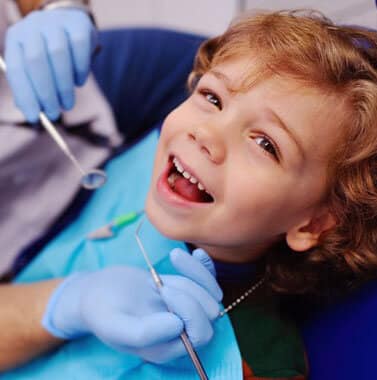 Young child smiling at orthodontist appointment while dentist holds mirror during dental examination.