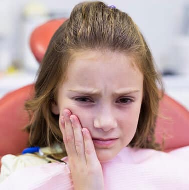 Young girl touching her cheek in discomfort, wearing pink shirt in clinical setting.