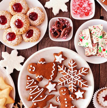 Assorted holiday cookies and treats arranged on white plates on wooden surface.