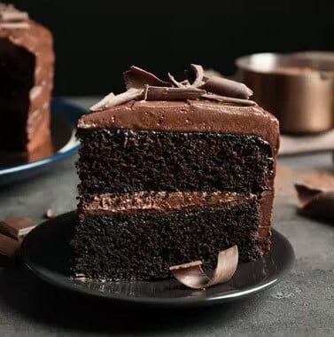 Decadent chocolate layer cake slice with frosting and chocolate shavings on dark plate.