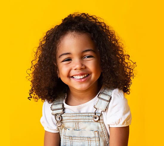 Smiling young child wearing overalls against bright yellow background for orthodontics website