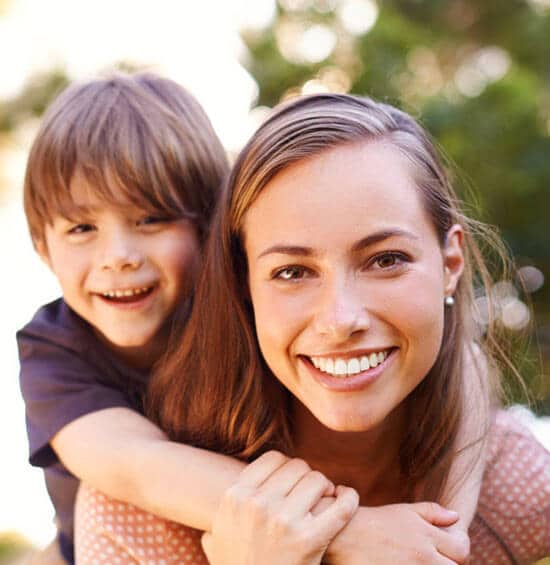 Woman and child smiling outdoors, embracing together in natural daylight setting.
