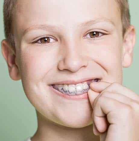 Young boy with braces smiling and pointing to teeth against green background.