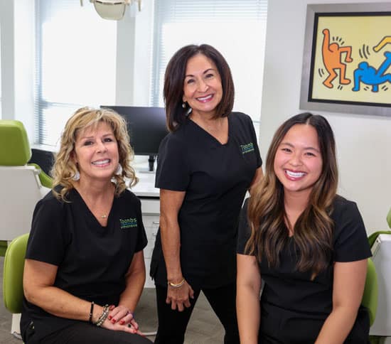 Three female orthodontic staff members in black uniforms smiling in a modern dental office.