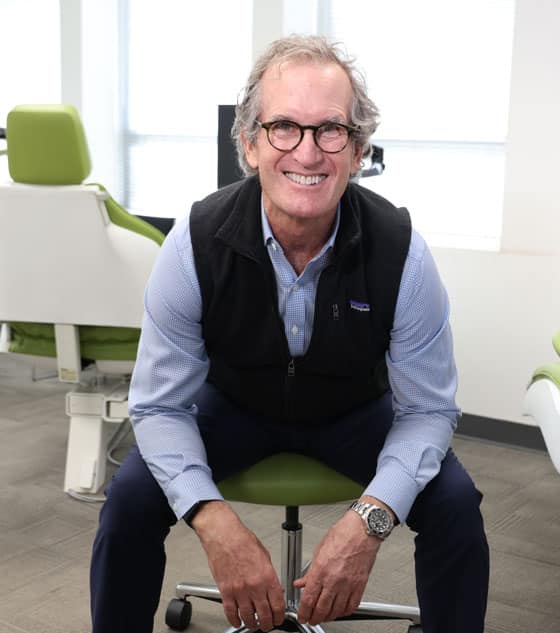 Professional man in office setting, wearing glasses and vest, seated on green chair, smiling at camera.
