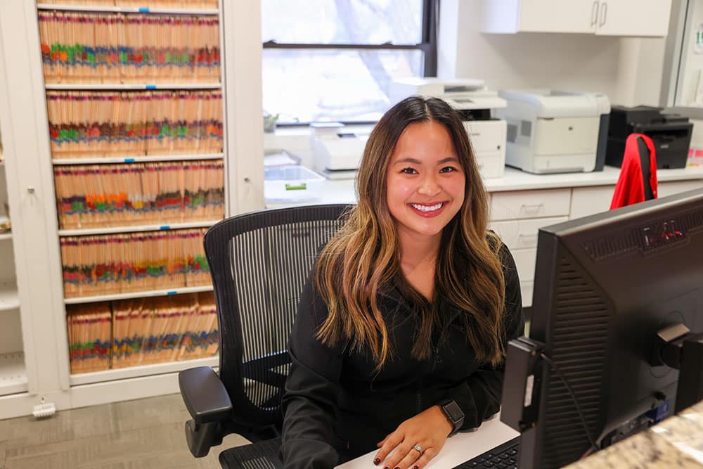 Woman smiling at desk in orthodontic office with law books visible in background.