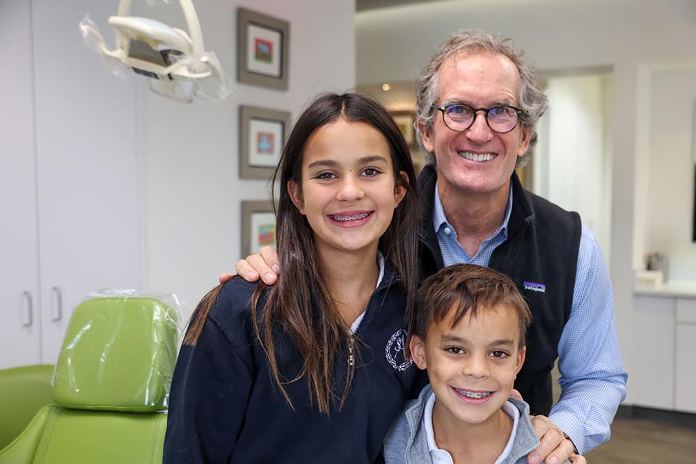 Man and two children smiling together in orthodontic office with green chair visible.