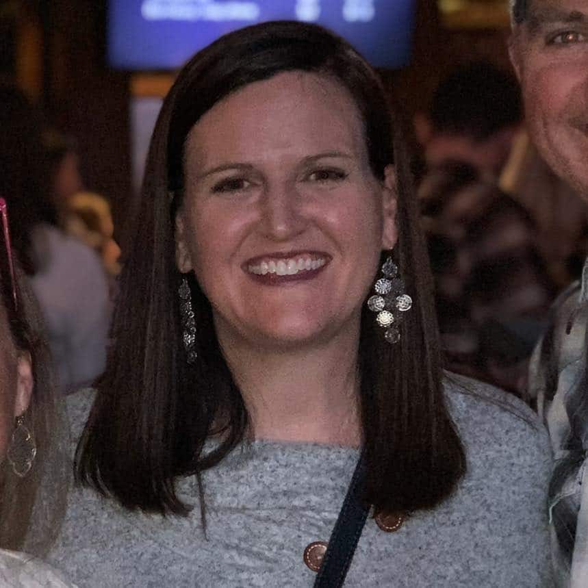 Woman smiling at social gathering, wearing pearl earrings and gray top, indoor event setting.