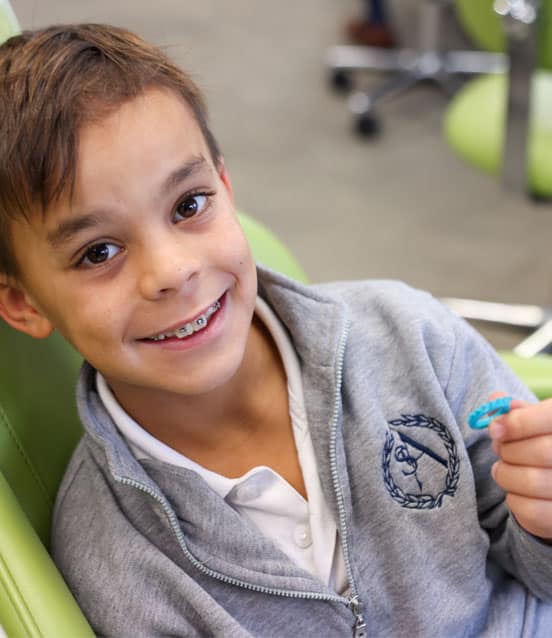 Young girl smiling at orthodontist's office, holding blue toothbrush, wearing gray hoodie.