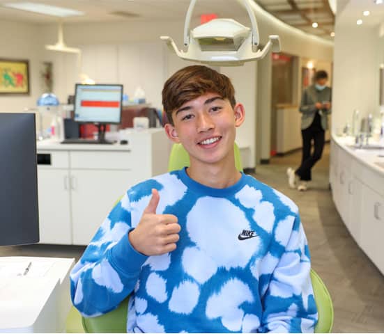 Young boy giving thumbs up in blue cloud-patterned shirt at orthodontics office reception area.