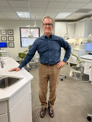 Man in blue shirt and tan pants standing in orthodontic office with dental equipment visible.