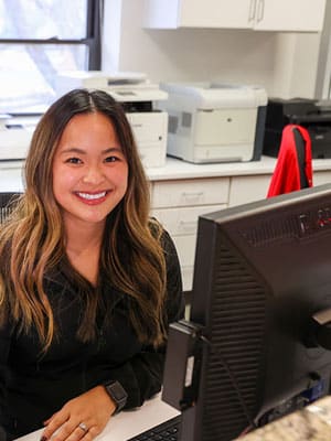 Smiling woman at desk with computer in modern orthodontic office setting.