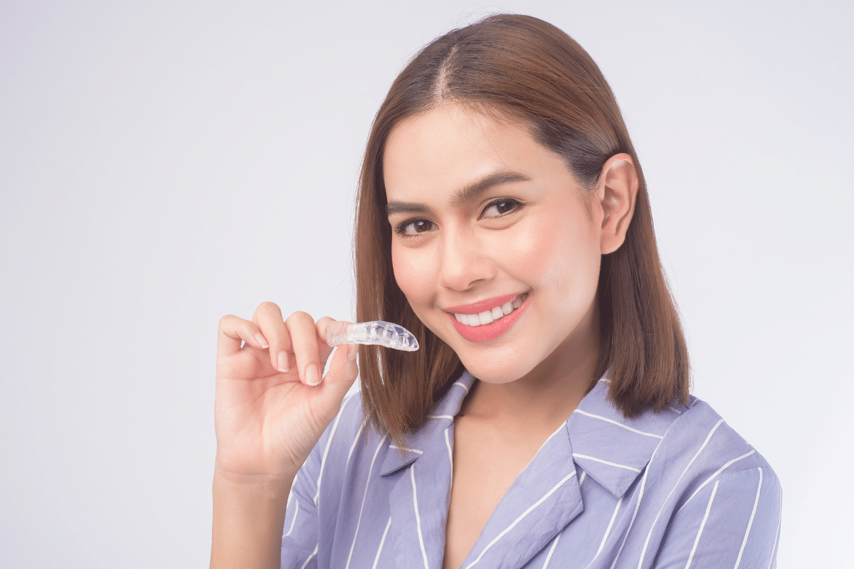 Young woman holding an Invisalign clear aligner tray, smiling to show teeth whitening results.