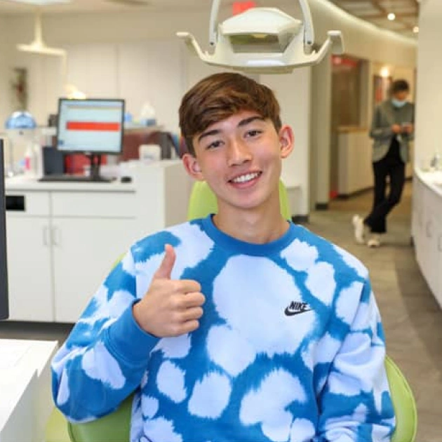 Young boy giving thumbs up in blue patterned shirt at orthodontics clinic reception area.