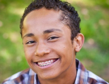 Young person smiling at camera outdoors with blurred green background, wearing plaid shirt.