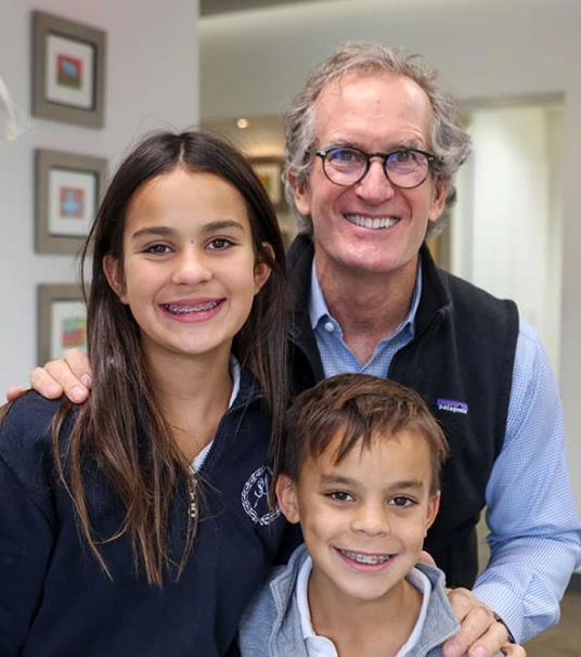 Man and two children smiling together in orthodontics office hallway with framed pictures on wall.
