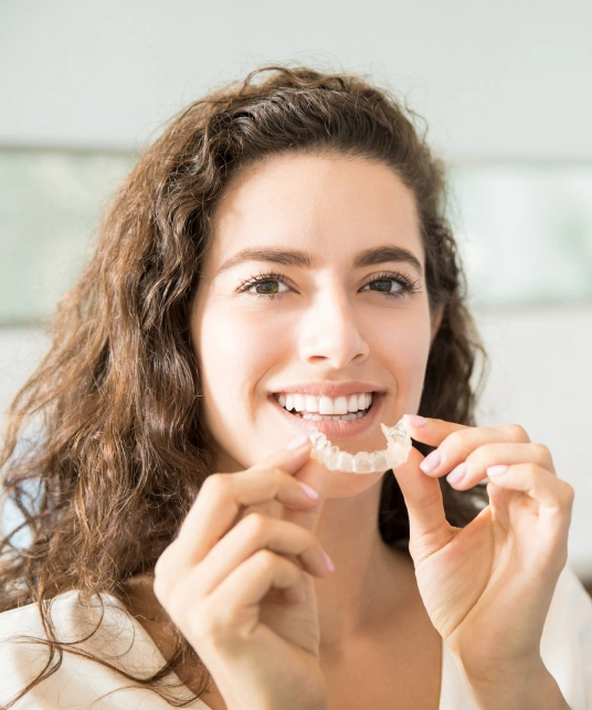 Woman inserting clear aligner tray, smiling in bright clinical setting.