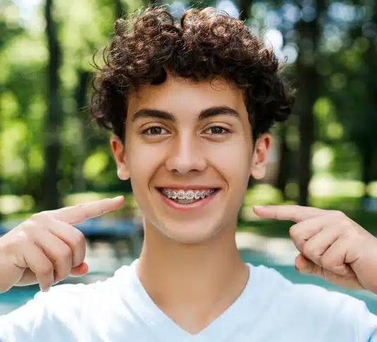 Smiling teenage boy holding clear aligners outdoors with green background.