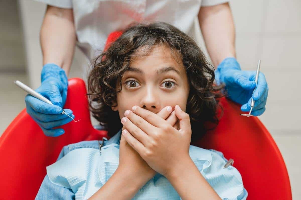 Child covering mouth with hands, scared during dental visit with dentist holding tools.